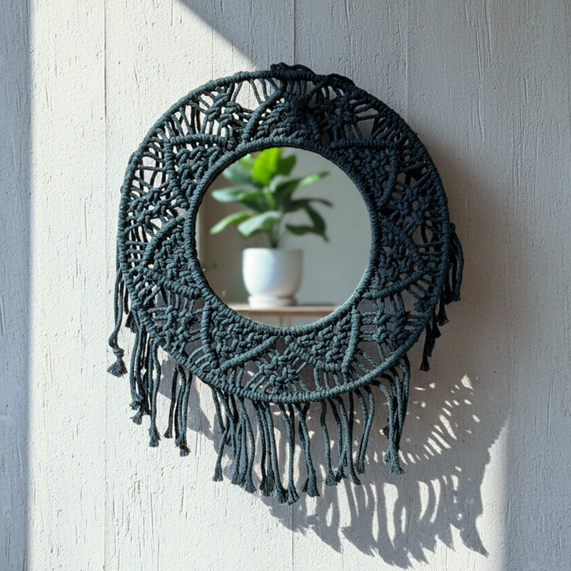 Round mirror with black macrame frame and fringe, reflecting a potted green plant