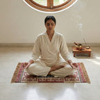 Person in white attire sitting in meditation on a patterned rug with a small table and incense holder in the background.