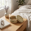 Wooden nightstand with a woven basket, book, glasses, and vase with lavender near a bed.