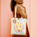 Woman holding a white tote bag with orange and yellow design against a pink background
