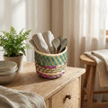 Woven basket with kitchen utensils on a wooden surface, with a plant and stool in the background.