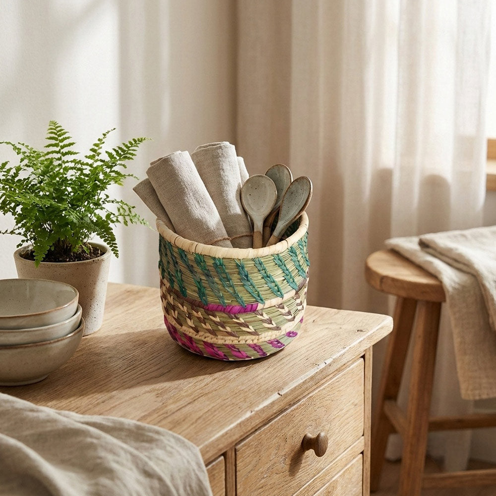 Woven basket with kitchen utensils on a wooden surface, with a plant and stool in the background.
