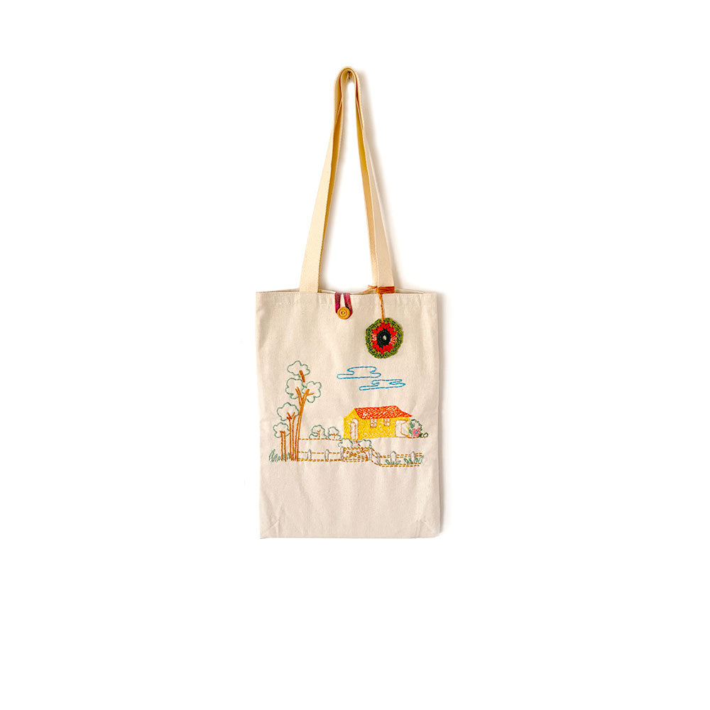 beige canvas tote bag with embroidery of house, trees, and sky, featuring a hanging flower ornament