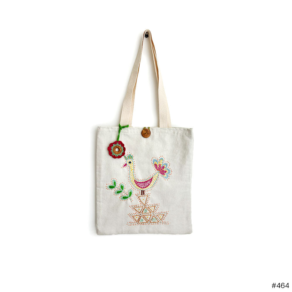 White canvas tote bag with colorful embroidered bird and geometric patterns, wooden button, and red flower charm