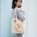 Woman holding a beige tote bag with floral embroidery against a light blue wall.