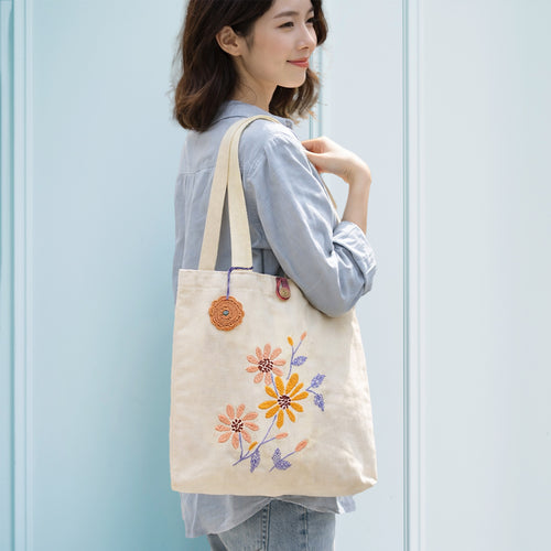 Woman holding a beige tote bag with floral embroidery against a light blue wall.