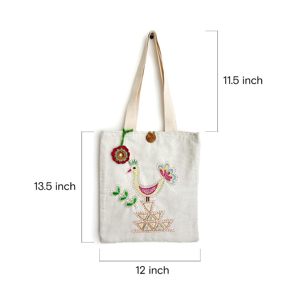 White canvas tote bag with colorful embroidered bird and geometric design, natural straps, wooden button, and flower charm