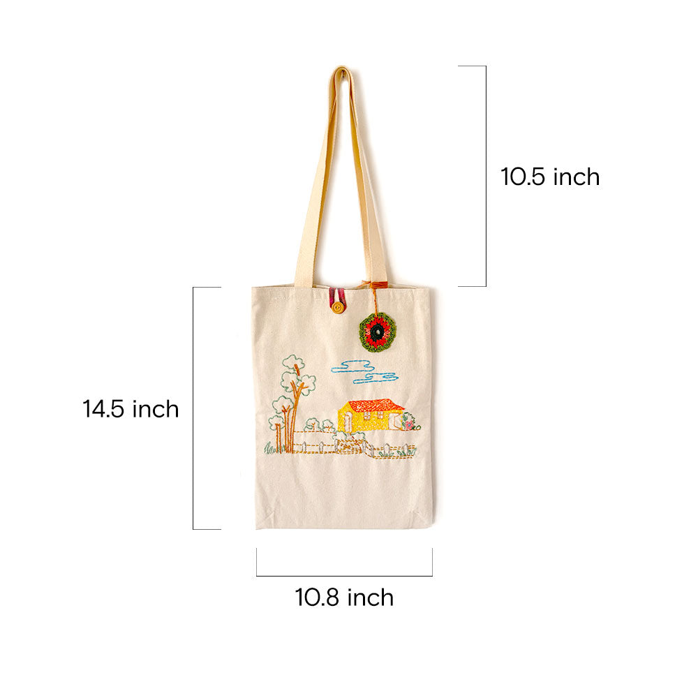 Beige canvas tote bag with embroidered house and trees design, dimensions labeled