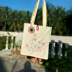 Beige canvas tote bag with colorful embroidered flowers and crochet keychain, outdoor sunny setting
