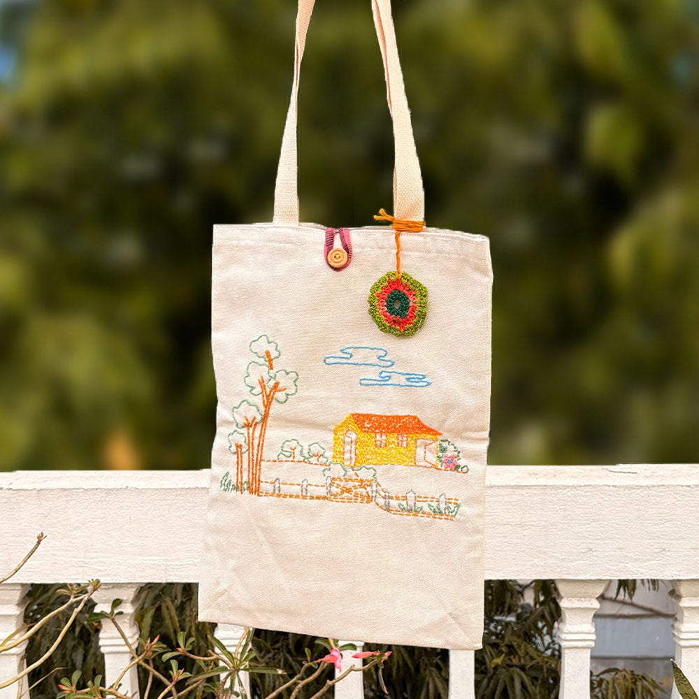 Beige canvas tote bag with embroidered house, trees, clouds, button and colorful crochet decoration