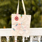 Beige canvas tote bag with embroidered pink bird, colorful feathers, and text 'feel pretty' hanging outdoors