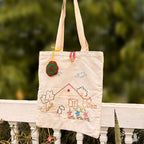 Beige canvas tote bag with embroidered house, flowers, trees, clouds, and a crocheted flower keychain