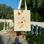Beige tote bag with floral embroidery held by a person outdoors.