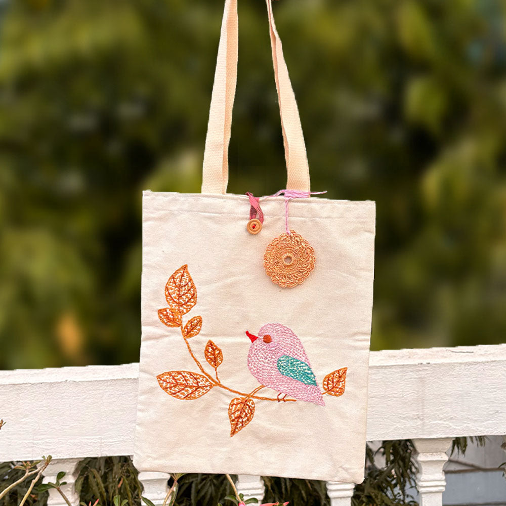 Cream tote bag with embroidered pink bird and orange leaves, hanging outdoors on white railing