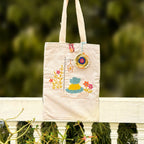 Beige fabric tote bag with colorful embroidered floral and dress design, hanging outdoors