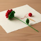 Crocheted red flower and green leaves on a wooden surface with a white paper background