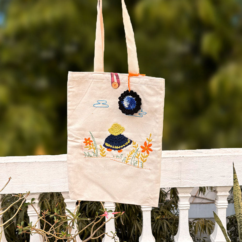 Beige canvas tote bag with embroidered girl, flowers, and clouds hanging on white railing outdoors
