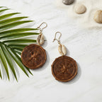 Pair of brown circular earrings with shell accents on a sandy background with palm leaf and stones.