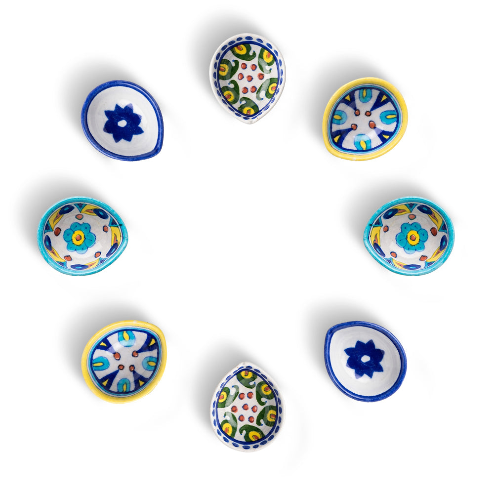 A top-down view of an assorted set of eight handcrafted blue pottery diyas featuring floral and geometric designs on a white background.