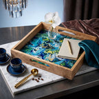 A square mango wood serving tray with blue and green marbleized enamel inlay sitting on a white marble surface, displaying a notebook, pen, and orchid.