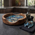 A round artisan mango wood serving tray with an enamel blue and green leaf print interior and sleek metal handles on a dark kitchen surface.