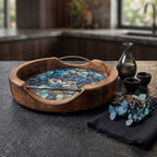 A round artisan mango wood serving tray with an enamel blue and green leaf print interior and sleek metal handles on a dark kitchen surface.