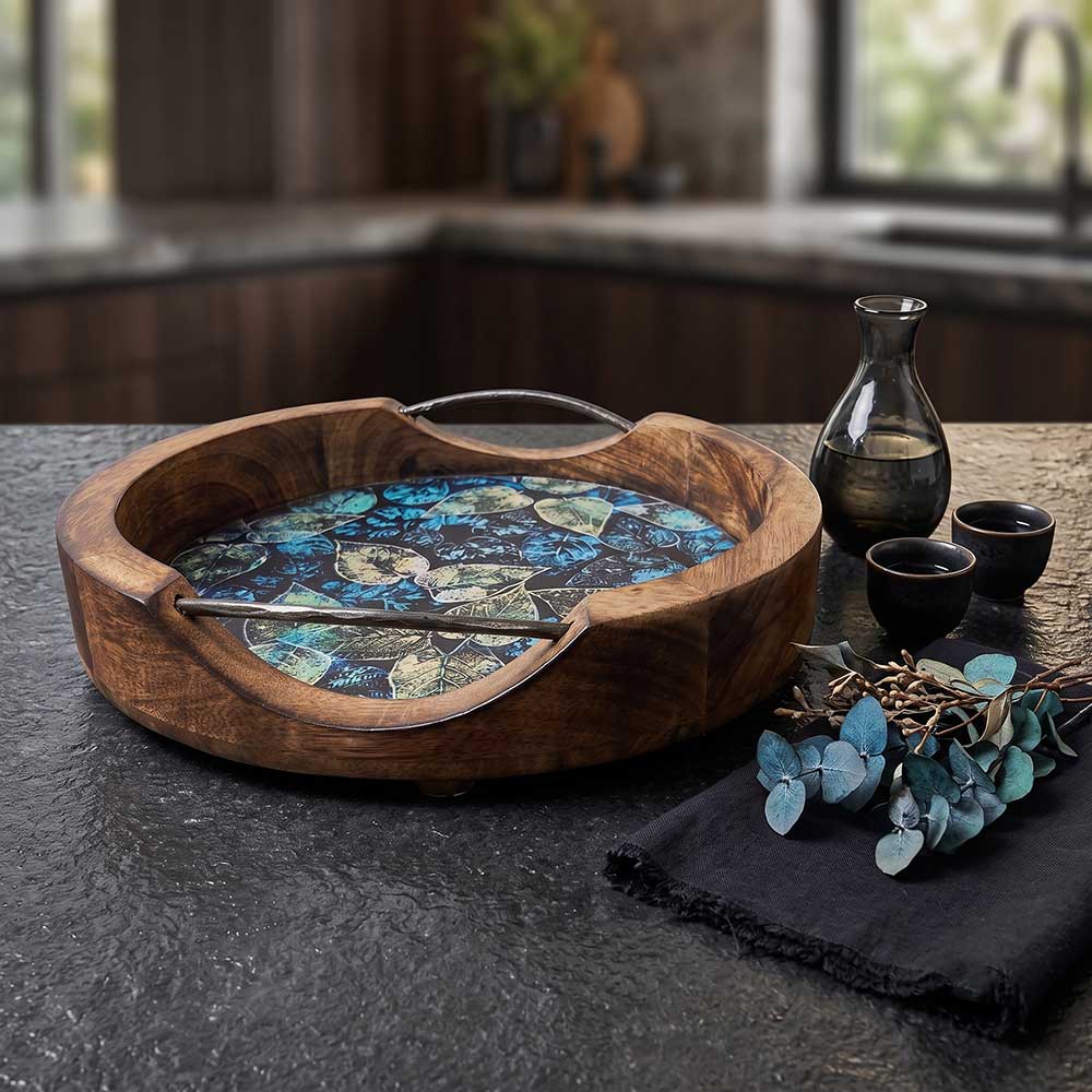 A round artisan mango wood serving tray with an enamel blue and green leaf print interior and sleek metal handles on a dark kitchen surface.