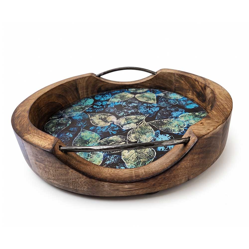Overhead view of a round mango wood serving tray showing intricate botanical leaf enamel print and metal handles.