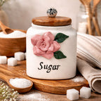 White sugar jar with wooden lid featuring a pink rose design on a wooden surface.
