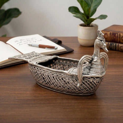 Decorative silver peacock-shaped box on a wooden surface with books and a plant in the background.